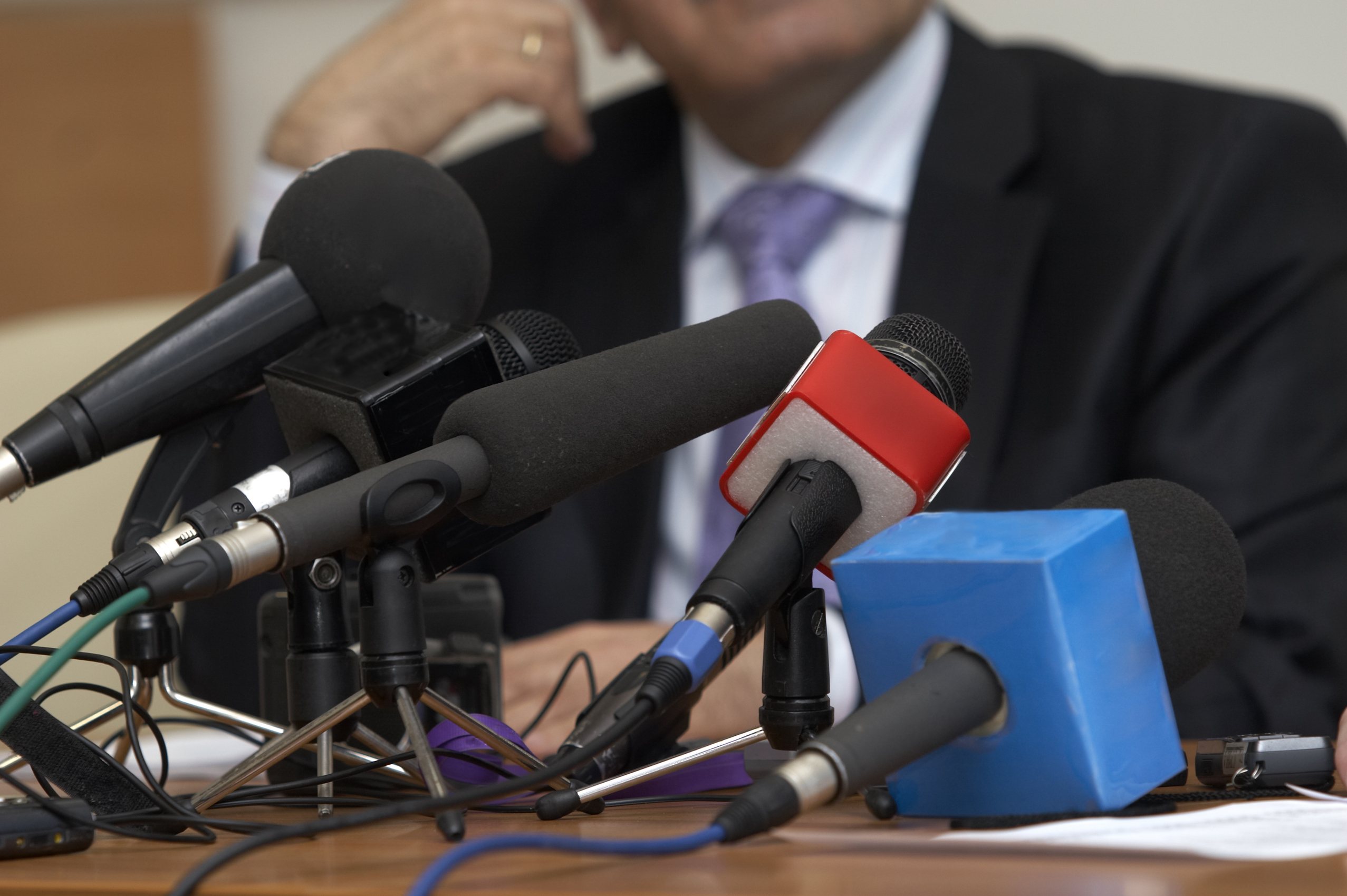 Spokesperson in a suit and tie behind a cluster of news microphones