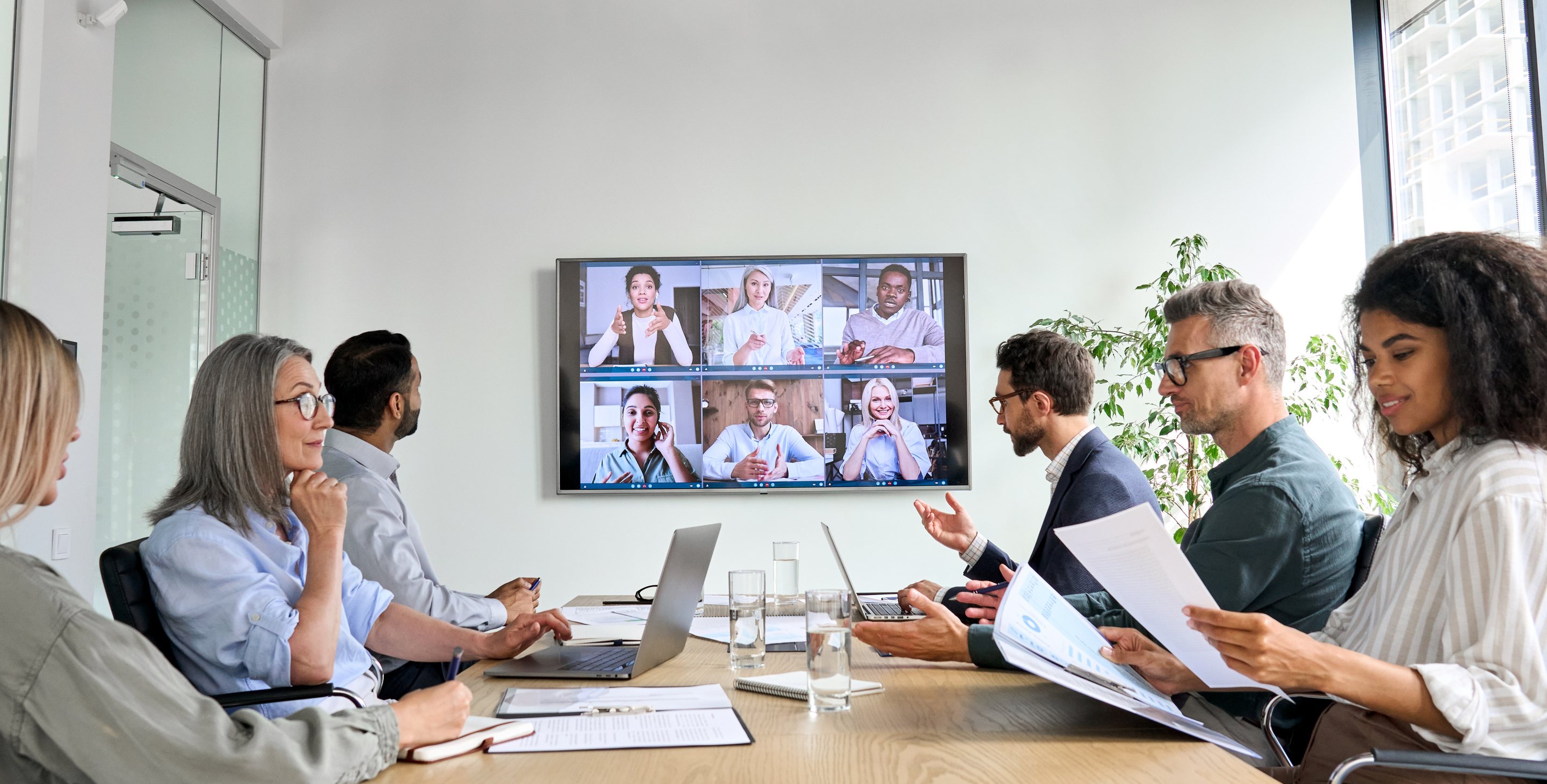 Executives at a conference table listening to colleagues on a video call screen